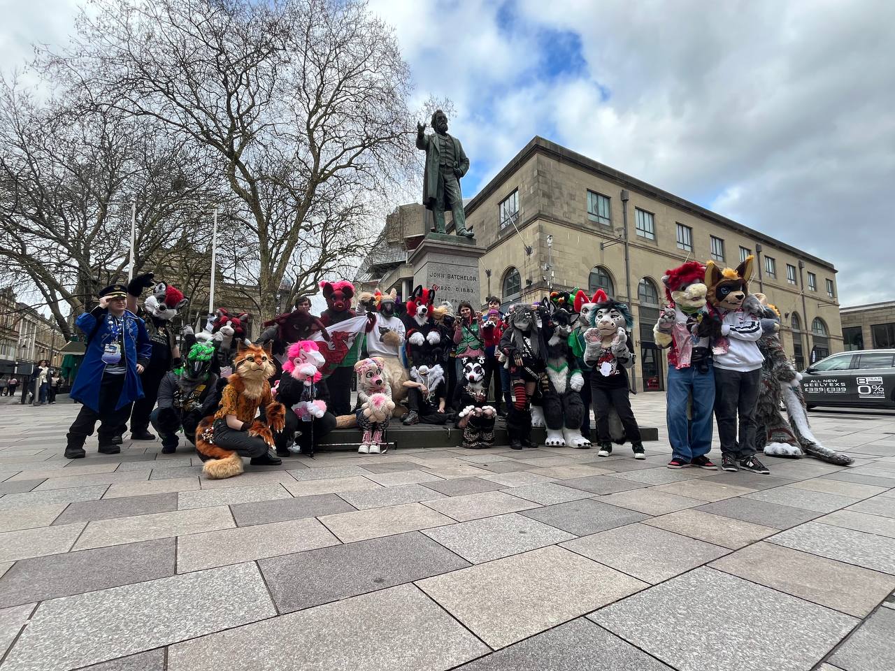 Photo of group of around 30 fursuiters together in front of a statue in Cardiff plaza outside